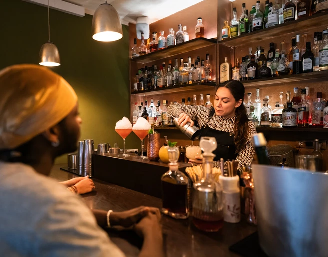 Toddy Tapper Eine Barkeeperin mixt Cocktails in einer atmosphärischen Bar vor aufmerksamem Publikum.A bartender mixes cocktails in an atmospheric bar in front of an attentive audience.