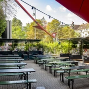 Zum Scheuen Reh Grüner Biergarten mit Holzbänken und Tischen, eingerahmt von Pflanzen, unter blauem Himmel.Green beer garden with wooden benches and tables, framed by plants, under a blue sky.