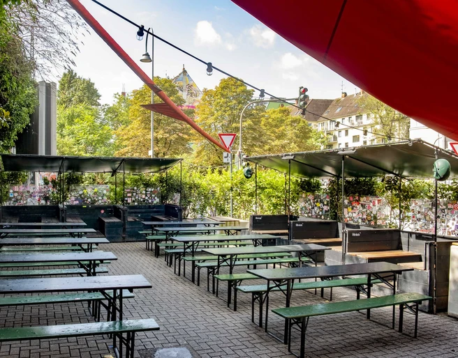 Zum Scheuen Reh Grüner Biergarten mit Holzbänken und Tischen, eingerahmt von Pflanzen, unter blauem Himmel.Green beer garden with wooden benches and tables, framed by plants, under a blue sky.