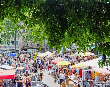 Flea market in Cologne-Nippes Eine lebendige Flohmarkt-Szene voller bunter Stände und emsiger Menschen unter schattenspendendem Laub.A lively flea market scene full of colorful stalls and busy people under shady foliage.
