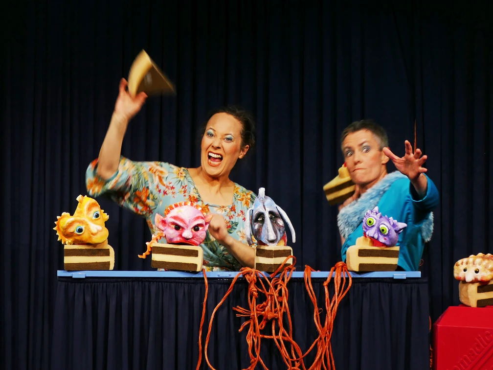 Knallwut Zwei Puppenspielerinnen animieren bunte, fantasievolle Puppen auf einer kleinen Bühne mit Vorhang.