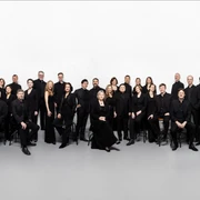 Rias Kammerchor Eine Gruppe von 28 Personen in schwarzer Kleidung posiert lächelnd in einem großen, weißen Raum.