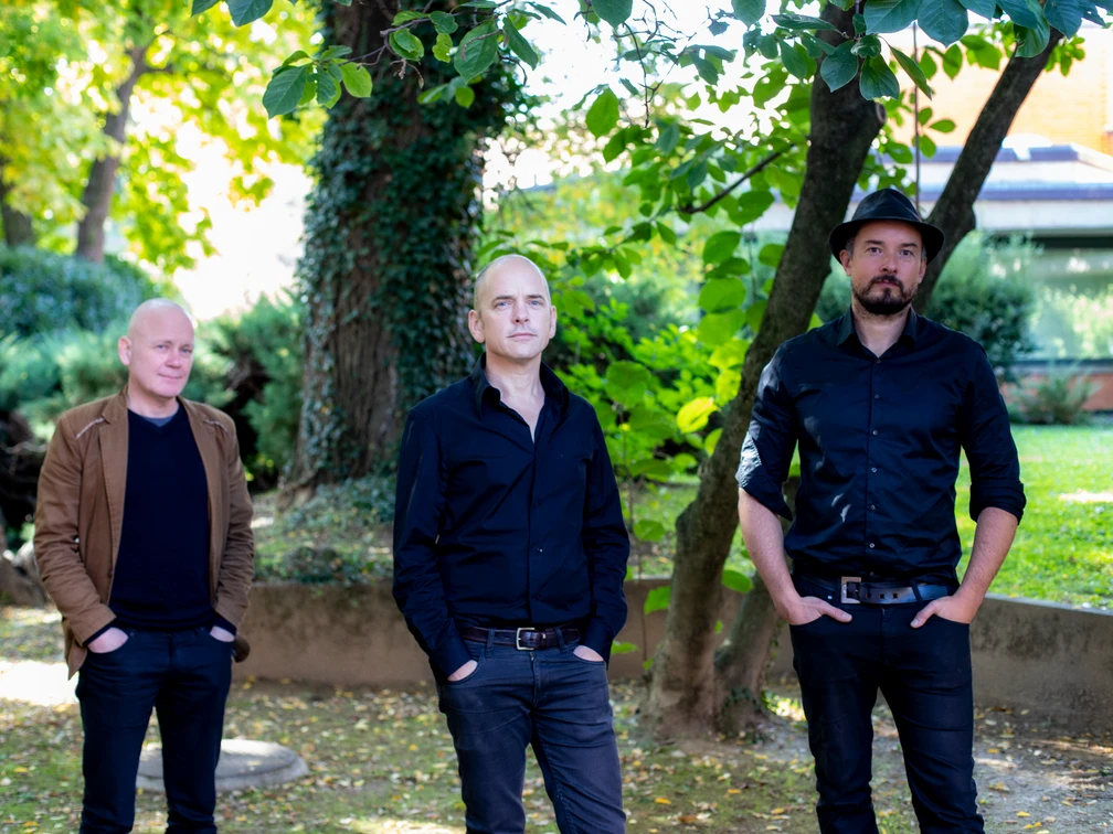 Tord Gustavsen Trio Drei Musiker posieren im Freien unter einem Baum; sie tragen dunkle Kleidung und blicken ernst.