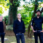 Tord Gustavsen Trio Drei Musiker posieren im Freien unter einem Baum; sie tragen dunkle Kleidung und blicken ernst.