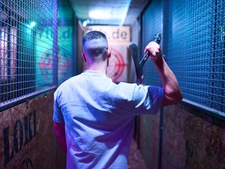 7th Space Cologne Ein Mann in einem Gang hält eine Axt, umgeben von Käfigwänden in violettem Licht.A man in a corridor holds an axe, surrounded by cage walls in violet light.