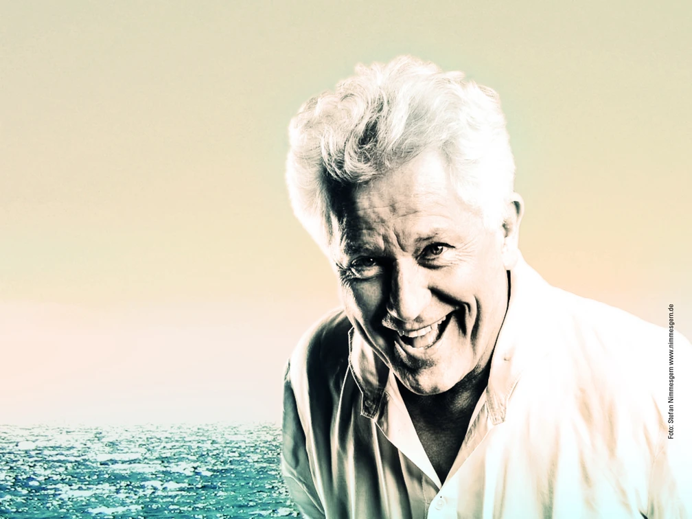 Alexis Sorbas Porträt eines lächelnden älteren Mannes vor einem weiten, sanft beleuchteten Ozean bei Sonnenuntergang.