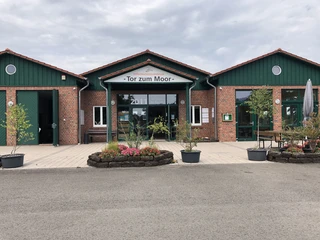 Infozentrum "Tor zum Moor" Backsteinbau mit grünem Dach und Glasfronten, umgeben von Pflanzen, mit Schild "Tor zum Moor".