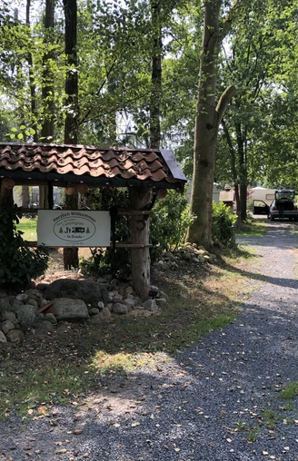 Eingang des Naturcampingplatzes "Im Braucke" mit Holzschild, umgeben von hohen Bäumen und Kiesweg.