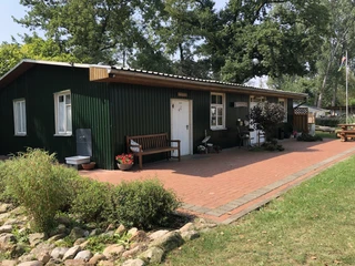Grünes Gebäude auf einem Campingplatz mit Terrasse, Bank, Pflanzen und umgeben von Bäumen.