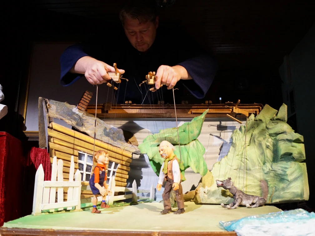 Spielszene, Peter Wolf  Eine Marionettenszene von Peter und Wolf vor einem rustikalen Haus, gelenkt von Puppenspielerhänden.