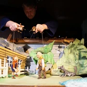 Spielszene, Peter Wolf  Eine Marionettenszene von Peter und Wolf vor einem rustikalen Haus, gelenkt von Puppenspielerhänden.