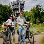 Am Baumwipfelpfad in Bad Iburg Zwei Menschen fahren bei sonnigem Wetter mit E-Bikes vor einem Aussichtsturm im Grünen.