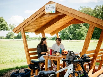 Picknickpause an der Friedensroute  Ein überdachter Holzpavillon auf einer Wiese mit zwei Personen und Fahrrädern im Vordergrund.