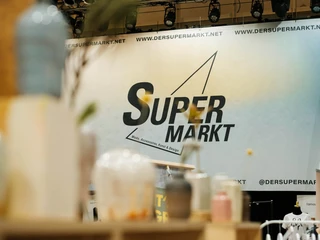 The Super Christmas Market Moderner und stilvoller Stand auf einem Weihnachtsmarkt. Im Vordergrund stehen stilvolle Glasgefäße auf einem Regal, während im Hintergrund eine große Werbewand mit dem Schriftzug "Super Markt" zu erkennen ist.Modern and stylish stand at a Christmas market. In the foreground are stylish glass jars on a shelf, while a large advertising wall with the words "Super Markt" can be seen in the background.