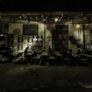 Gebäude 9 Bunte Lichterketten erhellen eine Außenfassade, während Menschen an rustikalen Holztischen sitzen.Colorful fairy lights illuminate an exterior façade while people sit at rustic wooden tables.