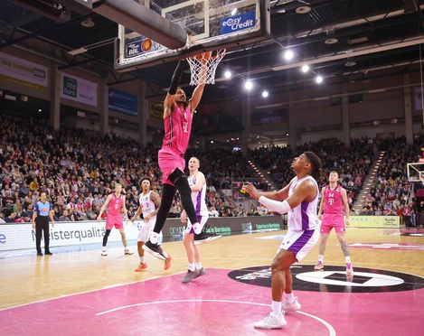 Telekom Baskets Bonn Spieler in pinken Trikots dominieren ein Basketballspiel in einer großen, voll besetzten Arena.Players in pink jerseys dominate a basketball game in a large, packed arena.