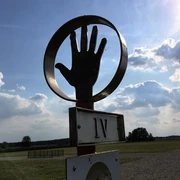 Römerlager Delbrück-Anreppen Skulptur einer Hand in einem Kreis vor einem weiten Feld, Wolken und blauem Himmel im Hintergrund.