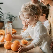 pexels-vlada-karpovich-4617285.jpg Ein begeistertes Kind greift nach Orangen, während Eltern im Hintergrund lächelnd zusehen.