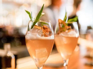 Suderman Zwei stilvoll arrangierte Cocktailgläser mit farbenfrohem Getränk und dekorativen Kräutern auf einer Holztheke.Two stylishly arranged cocktail glasses with a colorful drink and decorative herbs on a wooden counter.