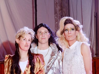 Drag & Drum Drei Personen in extravaganten Drag-Kostümen stehen selbstbewusst in einem bunt beleuchteten Bühnenraum.Three people in extravagant drag costumes stand confidently in a colorfully lit stage area.