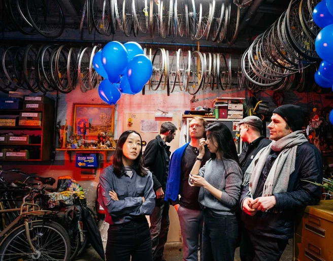 Maria de Alvear & hand werk In einem Raum mit vielen Fahrradrädern an der Decke hängen blaue Luftballons, umgeben von Menschen in entspannter Atmosphäre.Blue balloons hang from the ceiling in a room with lots of bicycles, surrounded by people in a relaxed atmosphere.