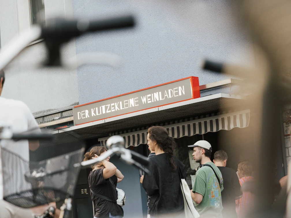 Der klitzekleine Weinladen Vor einem kleinen Geschäft in der Stadt versammeln sich Menschen. Fahrräder sind im Vordergrund.People gather in front of a small store in the city. Bicycles are in the foreground.