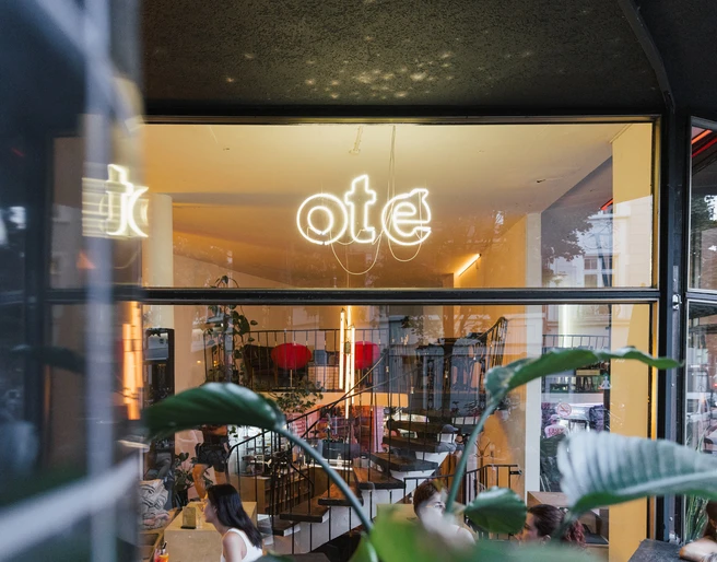 Oté Das Bild zeigt die helle und einladende Eingangsfassade eines Cafés oder Restaurants mit großen Glasfenstern und stilvoller Inneneinrichtung.