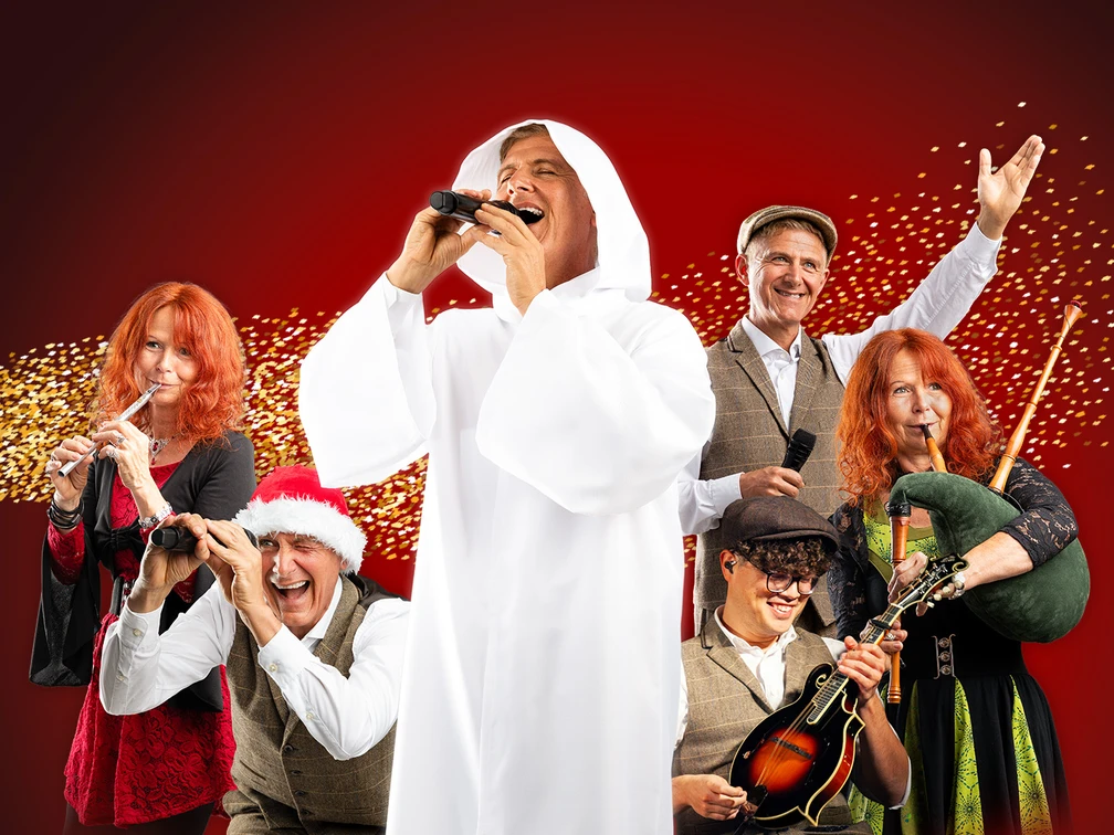 16x9_Hauptbild.jpg Eine fröhliche Gruppe von Musikern in festlicher Kleidung posiert mit Instrumenten auf rotem Hintergrund.