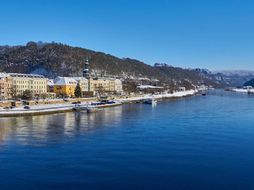 Blick auf Bad Schandau Flussufer mit pastellfarbenen Gebäuden, bewaldeten Hügeln und blauem Himmel in winterlicher Atmosphäre.