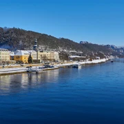 Blick auf Bad Schandau Flussufer mit pastellfarbenen Gebäuden, bewaldeten Hügeln und blauem Himmel in winterlicher Atmosphäre.
