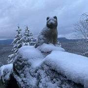 Katzstein Schnee bedeckte Katzenskulptur auf Felsen vor verschneiter Waldaussicht im Hintergrund.