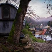Krippen Ein Baumhausfass befindet sich an einem Hang, im Hintergrund eine ruhige Dorfstraße mit bunten Häusern.
