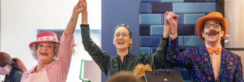 Arsen und Käsekuchen_schmal.jpeg Drei fröhliche Personen halten ihre Arme in die Luft und stehen nebeneinander vor blauen Fliesen.