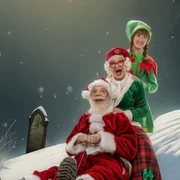 Mord am Nordpol_Schlitten.jpeg Zwei lachende Personen im Weihnachtskostüm sitzen auf einem verschneiten Hügel vor dunklem Hintergrund.