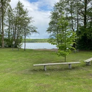 Badestelle Buchholz mit Bänken Eine große Wiese mit Bänken und Bäumen, im Hintergrund ein ruhiger See und bewölkter Himmel.