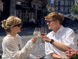 Basic 2 (c) Basic Gastrobar.JPG Ein junges Paar hebt lächelnd Gläser in einem sonnigen Straßencafé, umgeben von historischen Fassaden.