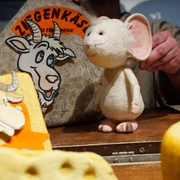 figurentheater-grinsekatze-liebe-gruesse-deine-tilda-1.jpg Eine weiße Stoffmaus auf einer Bühne zwischen großen Käseattrappen beim Figurentheater.