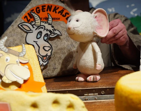 figurentheater-grinsekatze-liebe-gruesse-deine-tilda-1.jpg Eine weiße Stoffmaus auf einer Bühne zwischen großen Käseattrappen beim Figurentheater.