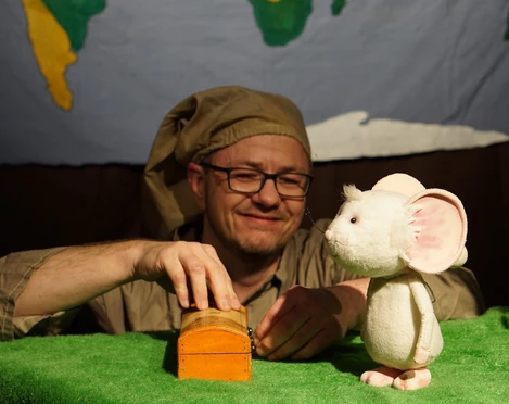 figurentheater-grinsekatze-liebe-gruesse-deine-tilda-2.jpg Puppenspieler mit Schlafmütze spielt auf grüner Bühne mit weißer Mausfigur und kleiner Holzkiste.