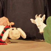 figurentheater-grinsekatze-das-letzte-schaf-1.jpg Ein Stoffschaf mit rot-grünem Schal steht vor einer gezeichneten Ziege hinter einer grünen Kulisse.