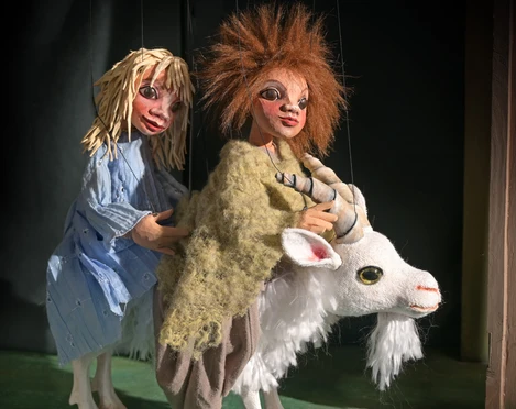 ambrella-figurentheater-zottelhaube-2.jpg Zwei bunte Marionetten reiten auf einer weißen Ziege in einer Theaterszene mit Bühnenlicht.