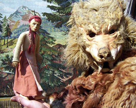 figurentheater-ernst-heiter-rotkaeppchen.jpg Holzfigur Rotkäppchen und wolfsähnliche Maske vor gemaltem Waldhintergrund einer Theaterbühne