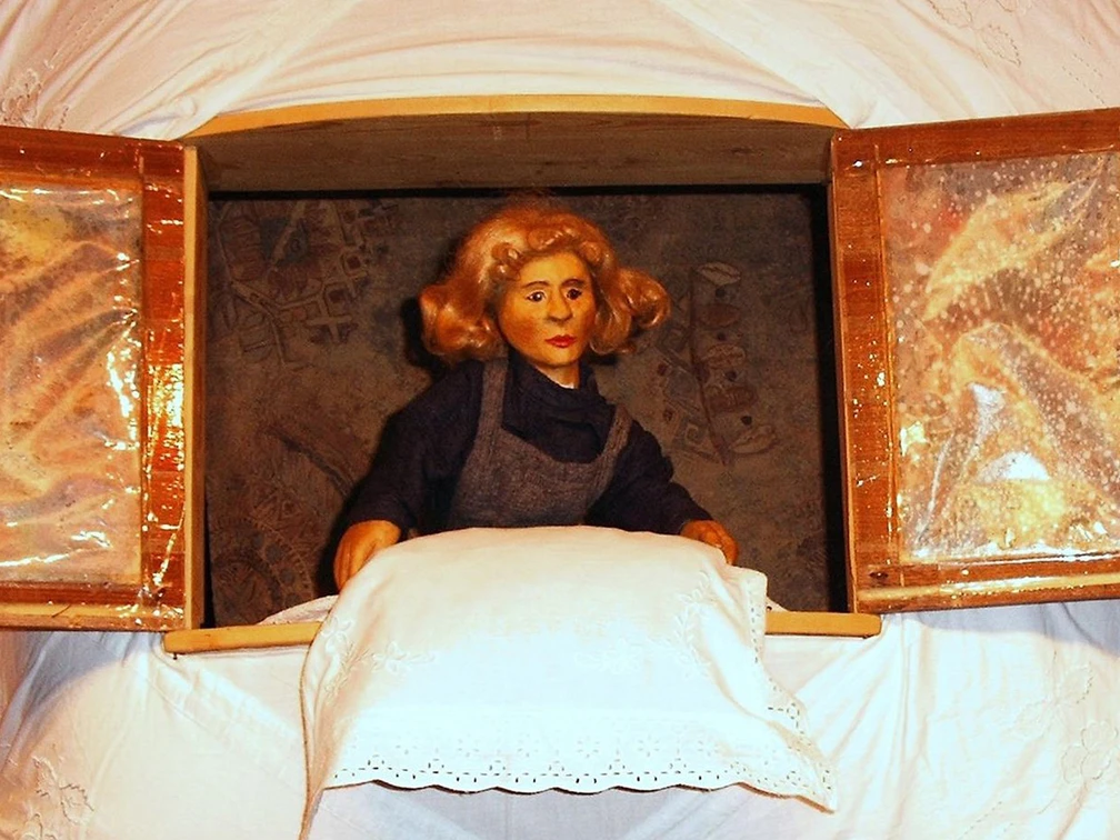 figurentheater-ernst-heiter-frau-holle.jpg Holzfigur der Frau Holle steht am Fenster und schüttelt ein weiß besticktes Kissen aus.