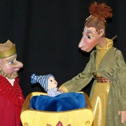 annes-buehne-der-prinz-mit-den-eselsohren-1.jpg Figurenpuppen eines Königs, einer Königin und eines Babys in Wiege vor dunklem Hintergrund