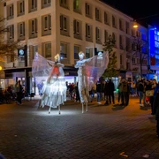 2024_11_02_Moonlight_Shopping_©HolgerBulk-169.jpg Zwei kostümierte Stelzenkünstler mit leuchtenden Flügeln erhellen eine Einkaufsstraße bei Nacht.Two costumed stilt artists with glowing wings light up a shopping street at night.Twee gekostumeerde steltenartiesten met gloeiende vleugels verlichten 's nachts een winkelstraat.