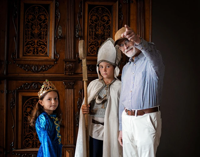 Online-Schloss-Kinderfuehrung-13.jpg Kinder in mittelalterlichen Kostümen und ein älterer Mann mit Hut in einem historischen Raum.