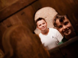 Online-Schloss-Kinderfuehrung-26.jpg Zwei Jungen blicken durch eine rustikale Holzstruktur und lachen in die Kamera.