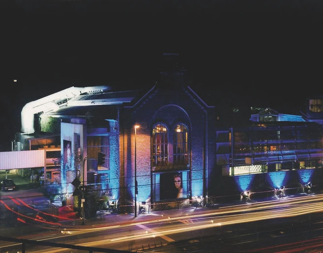 BALLONI Halle_1 Alte Backsteinhalle bei Nacht, beleuchtet in Blau, mit vorbeiziehenden Scheinwerferlichtern.Old brick hall at night, illuminated in blue, with passing spotlights.
