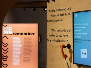View of the exhibition Eine moderne Ausstellungseinheit mit interaktivem Bildschirm und orangefarbenem Telefon, umgeben von thematischen Plakaten.A modern exhibition unit with an interactive screen and orange telephone, surrounded by themed posters.