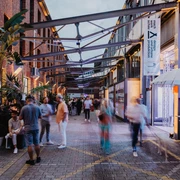 Menschen flanieren auf einem beleuchteten Innenhof mit roten Backsteingebäuden und tropischen Pflanzen.People stroll around an illuminated inner courtyard with red brick buildings and tropical plants.